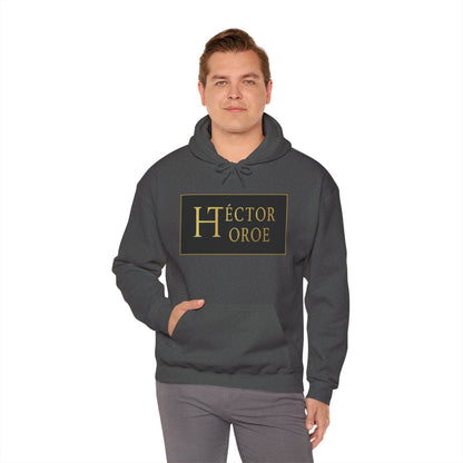 un hombre con sudadera brezo oscuro con capucha y bolsillo frontal, con un logotipo rectangular negro y dorado en el centro que muestra el texto “Héctor Toroe”.